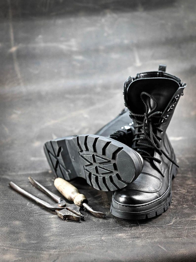 Puede incluir: Botas de cuero negro con cordones y una suela gruesa de goma. Las botas est&aacute;n sobre una superficie gris oscuro con un par de alicates y un destornillador.