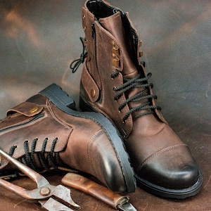 May include: Two brown leather boots with black laces and a zipper on the side. The boots are sitting on a brown leather surface with a pair of pliers and a small knife.