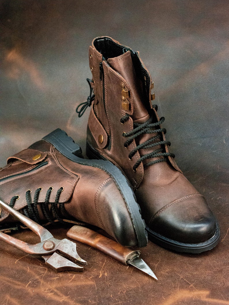 May include: Two brown leather boots with black laces and a zipper on the side. The boots are sitting on a brown leather surface with a pair of pliers and a small knife.