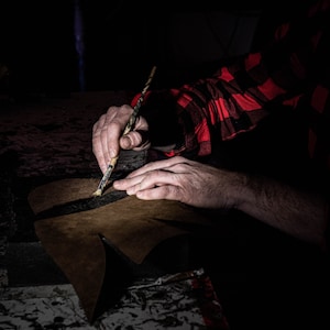 May include: A person's hands are shown working on a brown leather piece with a paintbrush. The person is wearing a red and black plaid shirt. The leather piece is on a dark surface.