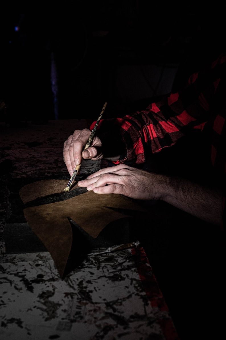 May include: A person's hands are shown working on a brown leather piece with a paintbrush. The person is wearing a red and black plaid shirt. The leather piece is on a dark surface.