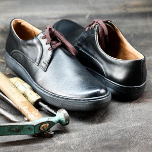 May include: Two black leather lace-up shoes with white soles. The shoes are on a gray surface with a wooden hammer and a metal tool.