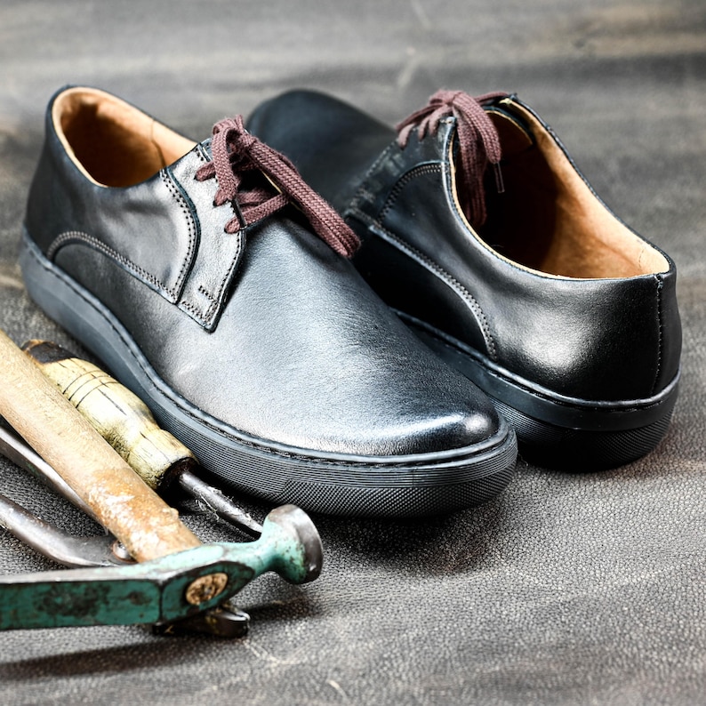 May include: Two black leather lace-up shoes with white soles. The shoes are on a gray surface with a wooden hammer and a metal tool.