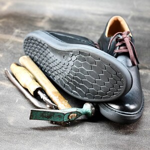 May include: A close-up of a black leather shoe with a textured sole. The shoe is lying on a brown surface next to two tools, one with a wooden handle and one with a green handle.