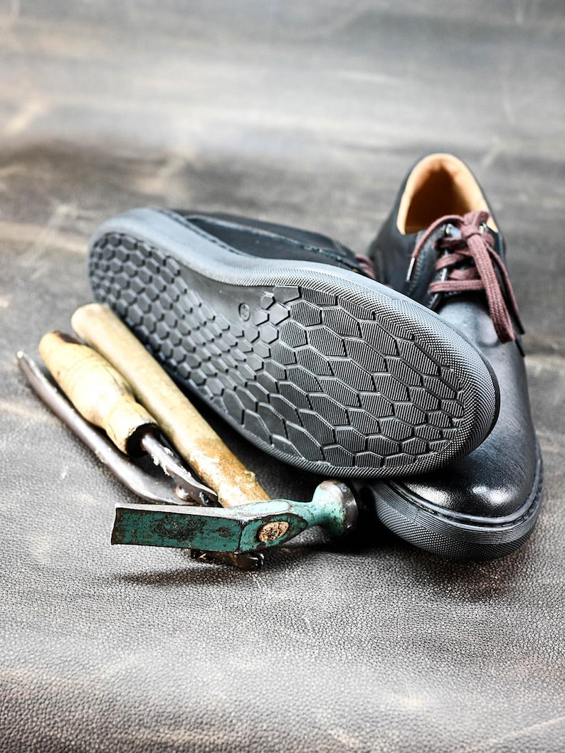 May include: A close-up of a black leather shoe with a textured sole. The shoe is lying on a brown surface next to two tools, one with a wooden handle and one with a green handle.