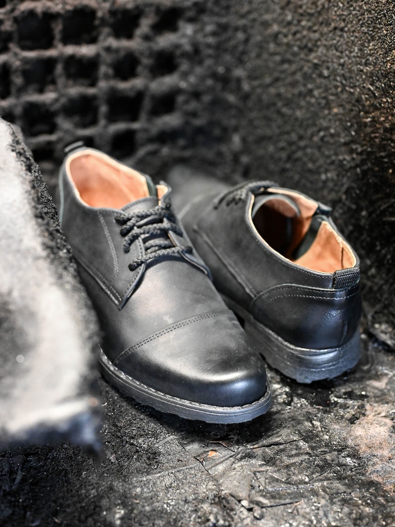 May include: Two black leather lace-up shoes with brown leather lining. The shoes are sitting on a dark, textured surface.