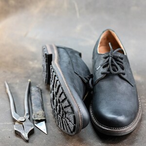 May include: Two black leather shoes with laces, one with a visible sole, and two tools for shoe repair: a pair of pliers and a utility knife.