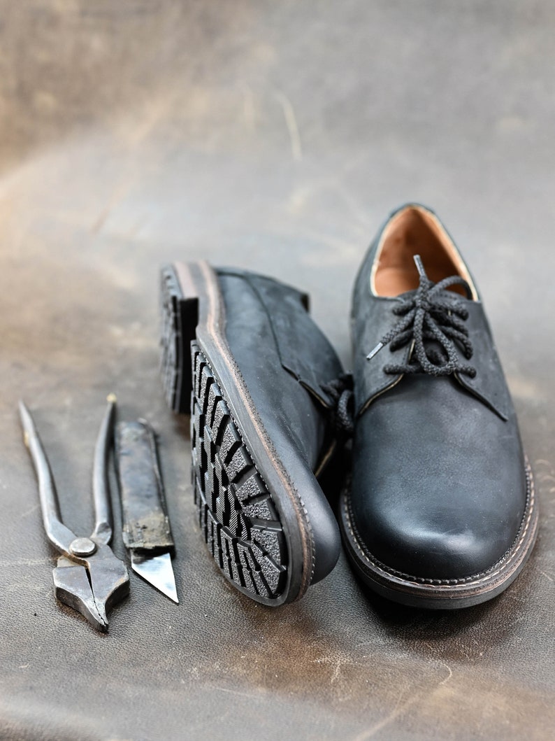 May include: Two black leather shoes with laces, one with a visible sole, and two tools for shoe repair: a pair of pliers and a utility knife.
