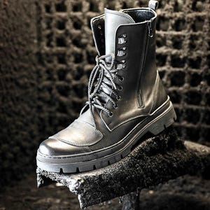 Puede incluir: Botas negras de cuero con cremallera lateral y cordones en la parte delantera. Las botas tienen una suela gruesa y est&aacute;n sobre una superficie rugosa y oscura.