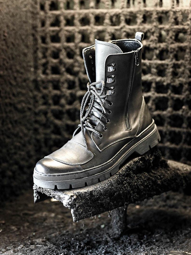 Puede incluir: Botas negras de cuero con cremallera lateral y cordones en la parte delantera. Las botas tienen una suela gruesa y est&aacute;n sobre una superficie rugosa y oscura.
