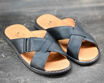 Op de afbeelding: Een paar zwarte leren sandalen met een kruisbandontwerp. De sandalen hebben een bruin voetbed en een donkergrijze zool. De sandalen worden gepresenteerd op een donkergrijze ondergrond.