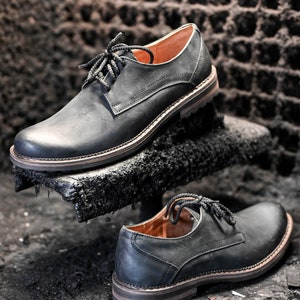 May include: Two pairs of black leather lace-up shoes with a classic design. The shoes have a slightly distressed look and are sitting on a dark surface.
