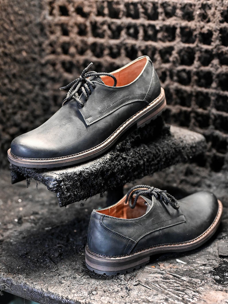 May include: Two pairs of black leather lace-up shoes with a classic design. The shoes have a slightly distressed look and are sitting on a dark surface.