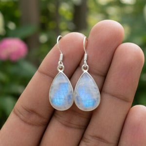 May include: Silver teardrop earrings featuring iridescent moonstone. The earrings have a simple hook design. The earrings are held against a blurred background of green foliage and flowers.