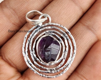 May include: A silver pendant with a purple amethyst gemstone set in a circular design with concentric circles. The pendant is on a hand.