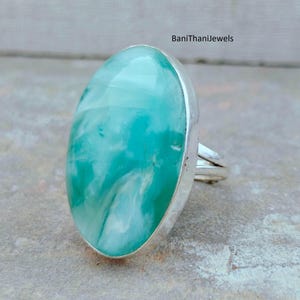 May include: A large, oval-shaped ring featuring a vibrant turquoise gemstone with white marbling, set in a silver band. The ring's design is simple, highlighting the stone's natural beauty. The ring is a statement piece.