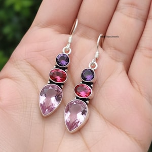 May include: A pair of silver dangle earrings featuring a teardrop-shaped, light pink gemstone at the bottom, a red oval stone in the middle, and a purple round stone at the top. The earrings are displayed on a hand.