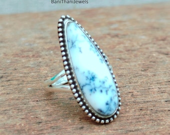 Puede incluir: Anillo de plata con una piedra grande en forma de lágrima, con base blanca y patrones dendríticos azul-gris. La piedra está enmarcada por un borde plateado con cuentas. Colocado sobre una superficie de madera.