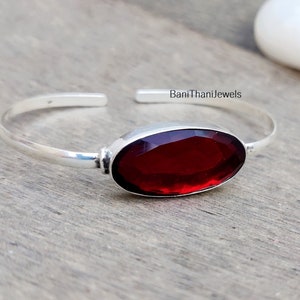Puede incluir: Un brazalete de plata con una gran piedra roja de forma ovalada. El brazalete está abierto y tiene un diseño sencillo.