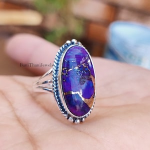 May include: A silver ring with a large oval purple and gold gemstone. The ring has a decorative border around the stone.
