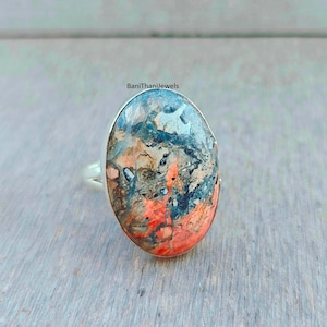 Puede incluir: Anillo ovalado de piedra preciosa con banda plateada. La piedra presenta una mezcla de colores rojo, azul y marrón. El anillo está sobre una superficie de madera gris claro. El texto "BaniThaniJewels" es visible.