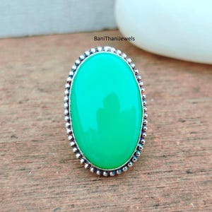 以下が含まれることがあります： 鮮やかな緑色の宝石をあしらった楕円形のリング。宝石は、ビーズの縁が付いた銀色の金属バンドにセットされています。リングは木製の表面に表示されています。ジュエリーです。