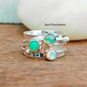 May include: A silver spinner ring featuring three opal gemstones in green and white hues. The ring has multiple bands and small silver beads. The text "BaniThanijewels" is visible.