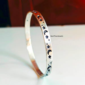 Puede incluir: Una pulsera de plata con un diseño celestial. La pulsera presenta un patrón repetido de lunas crecientes, estrellas y puntos. El texto "BaniThanijewels" es visible en el lado derecho de la imagen.