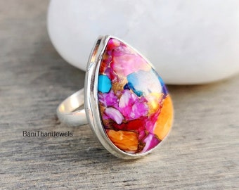 Puede incluir: Un anillo en forma de lágrima con una piedra preciosa multifacética de colores engastada en una banda de plata. La piedra preciosa presenta tonos de rosa, naranja, turquesa y oro.