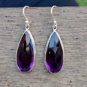 May include: A pair of teardrop-shaped earrings with faceted, deep purple gemstones set in silver-colored metal. The earrings have a hook closure. The brand name "BaniThaniJewels" is printed on the background.