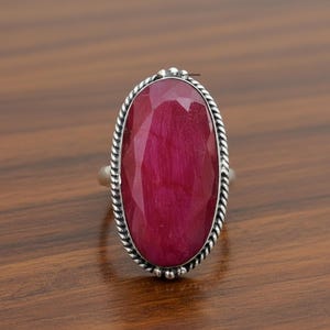 May include: A silver ring featuring a large, oval-cut ruby gemstone. The ruby is a deep red color and is surrounded by a twisted silver rope design. The ring is set on a wooden surface.
