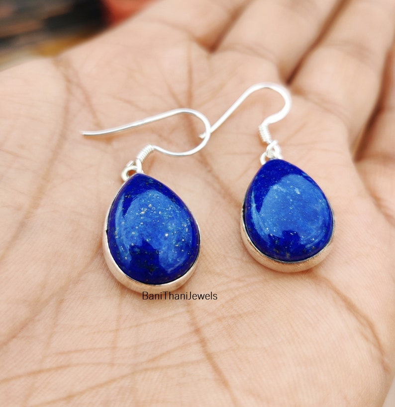 May include: A pair of silver dangle earrings featuring teardrop-shaped lapis lazuli stones. The stones are a deep blue colour with flecks of gold.