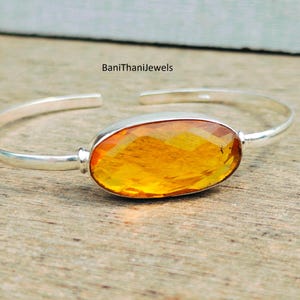 Op de afbeelding: Een zilveren armband met een grote, ovale amber edelsteen. De edelsteen is gefacetteerd en heeft een warme, gouden tint.