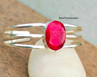 May include: A silver ring with a red oval gemstone set in the centre. The ring has three bands that wrap around the finger.