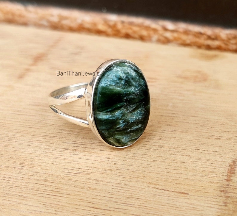 May include: A silver ring with a green and blue oval gemstone. The ring has a split band design. "BaniThaniJewels" is printed on the band.