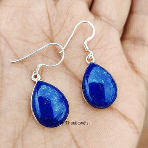 May include: A pair of silver dangle earrings featuring teardrop-shaped lapis lazuli stones. The stones are a deep blue colour with flecks of gold.