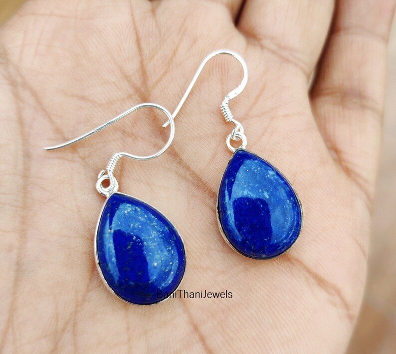 May include: A pair of silver dangle earrings featuring teardrop-shaped lapis lazuli stones. The stones are a deep blue colour with flecks of gold.