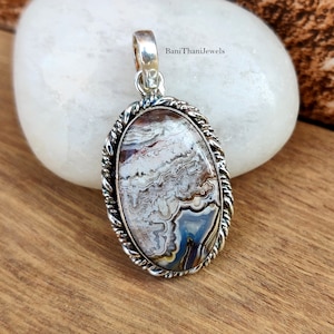 Puede incluir: Un colgante de plata con una piedra de ágata blanca, marrón y azul engastada en un borde de cuerda. El colgante tiene forma ovalada y tiene una anilla de plata.