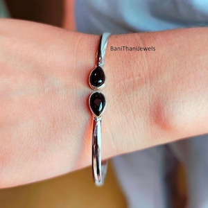 May include: A silver bracelet with two black teardrop-shaped stones. The bracelet is open-ended and sits on a wrist. The text "BaniThanijewels" is visible on the wrist.