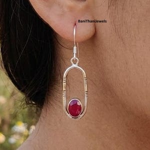 Peut inclure: Boucle d'oreille argentée avec une pierre précieuse rouge ovale. La boucle d'oreille a une forme ovale allongée et ouverte avec des détails gravés. Elle est suspendue à un crochet. Le texte "BaniThaniJewels" est visible en arrière-plan.