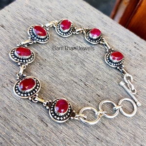以下が含まれることがあります： 赤い宝石のアクセントが付いたシルバーブレスレット。ブレスレットはトグル留め具が付いており、小さなシルバービーズで飾られています。木製の表面に「BaniThaniJewels」と印刷されています。