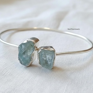 Puede incluir: Un brazalete de plata con dos piedras de aguamarina en bruto, de color azul verdoso pálido. Las piedras están engarzadas en plata y son el punto central del diseño minimalista. El brazalete se exhibe sobre una superficie blanca.