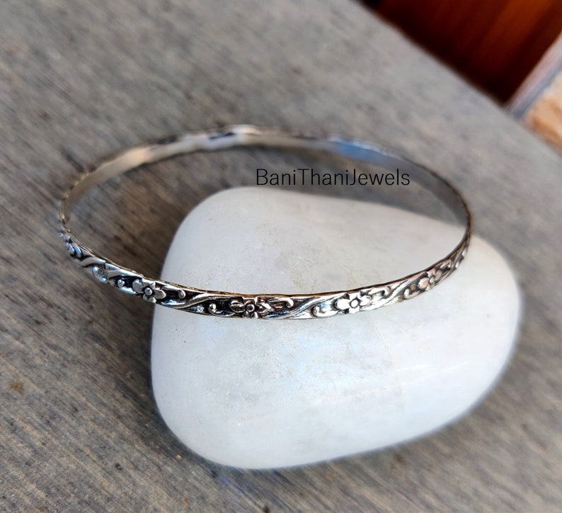 May include: A silver bangle bracelet featuring a delicate floral design. The bracelet is thin and has a simple, elegant look.
