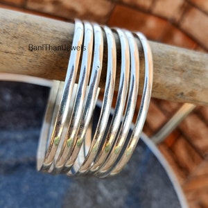 May include: A set of seven silver bangles stacked on a wooden surface. The bangles are smooth and shiny, and they are all the same width.
