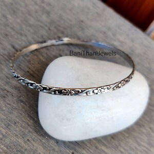 May include: A silver bangle bracelet featuring a delicate floral design. The bracelet is thin and has a simple, elegant look.