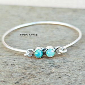 Puede incluir: Un brazalete de plata con dos piedras de ópalo azul claro. El brazalete está abierto y tiene un diseño sencillo.