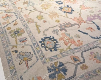 May include: A large, rectangular area rug with a cream-colored background and a floral pattern in shades of blue, peach, yellow, and purple. The rug has a detailed border with geometric and floral elements, adding to its traditional design.