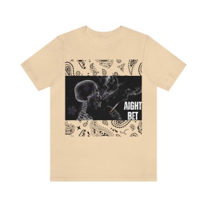 May include: A light beige t-shirt with a black and white graphic of a skeleton smoking a cigarette. The graphic is surrounded by a black paisley pattern. The text "AIGHT BET" is printed in white on the graphic.