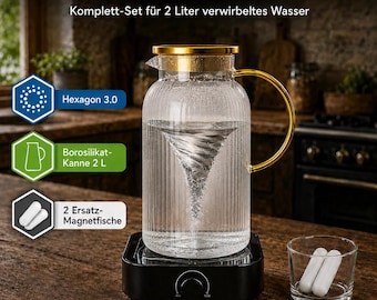 Può includere: Una caraffa in vetro scanalato trasparente con coperchio e manico dorati, posta su una base magnetica nera che crea un vortice d'acqua. Il set include due barrette magnetiche bianche in un bicchiere. Testo: HEXAGON 3.0 WASSERSET.