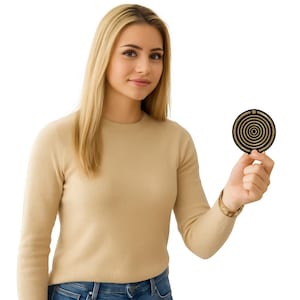 May include: A black and gold circular pendant with a spiral design is held up. The pendant has a small hole at the top. The person is wearing a tan sweater and blue jeans.